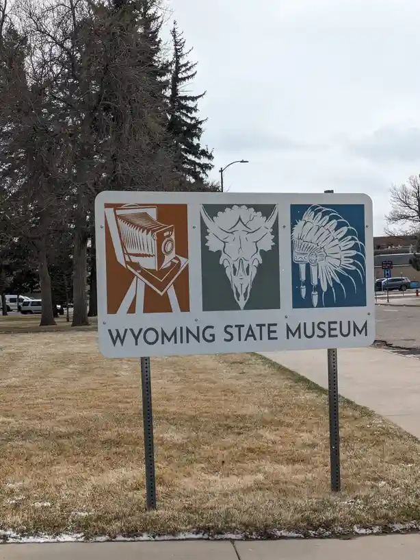 A white sign for the Wyoming State Museum displays three icons: a vintage camera, a bison skull, and a Native American…