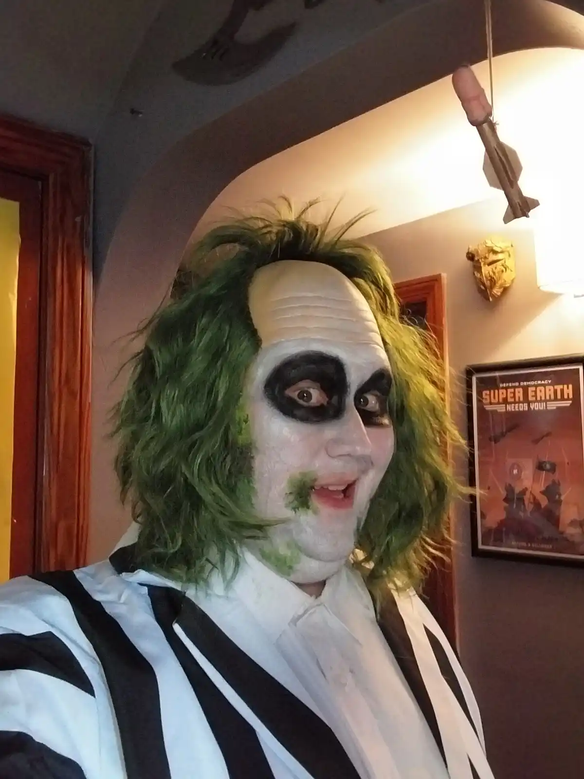 A person dressed as Beetlejuice with green hair, white face paint, and black eye makeup, wearing a striped shirt, looks at…