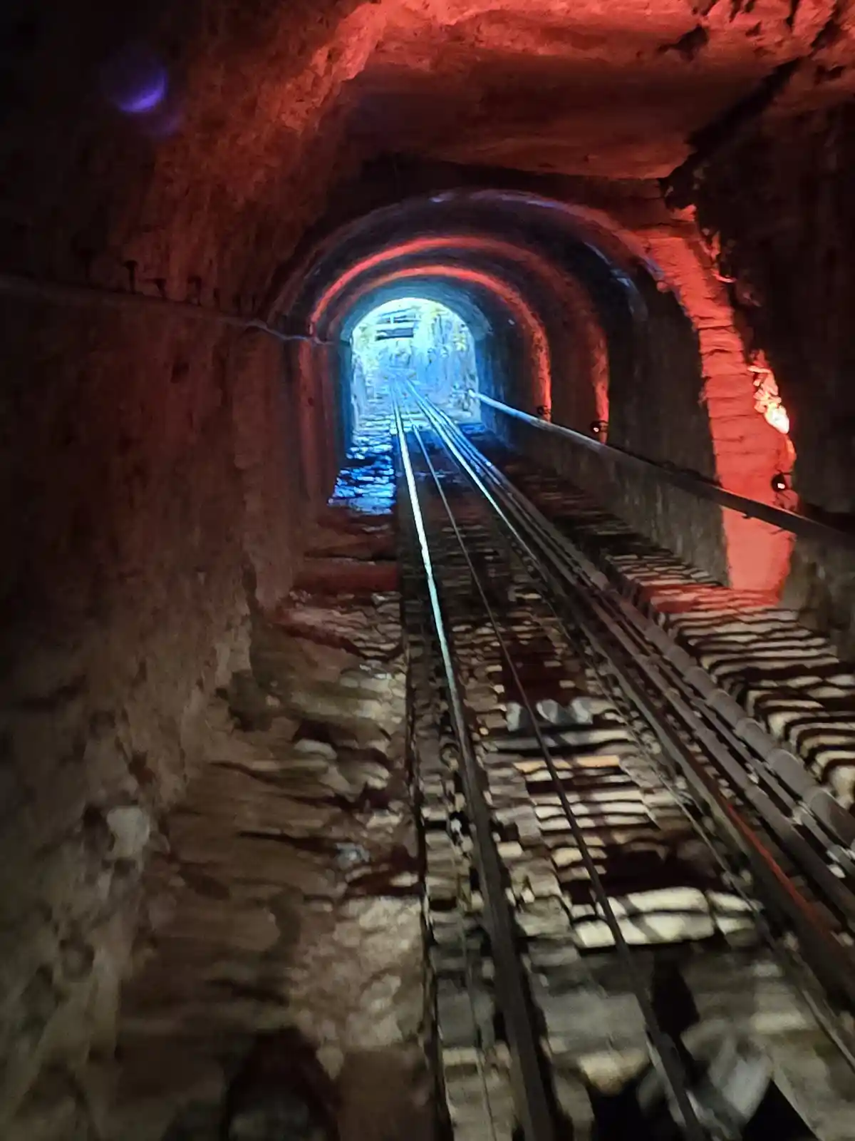 A view looking down a narrow, arched tunnel with railway tracks running toward a bright exit. The tunnel walls are rough…