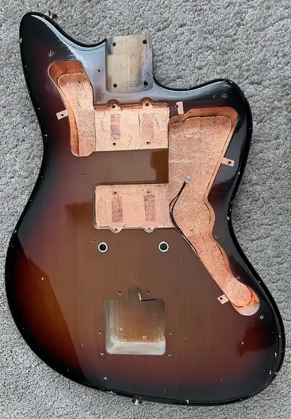 The image shows the back of a sunburst-finished electric guitar body lying on a gray carpet, with its pickguard and…