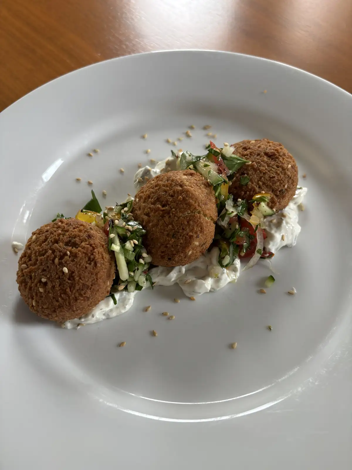 A white plate holds three golden-brown falafel balls resting on a bed of creamy white sauce, topped with chopped green…