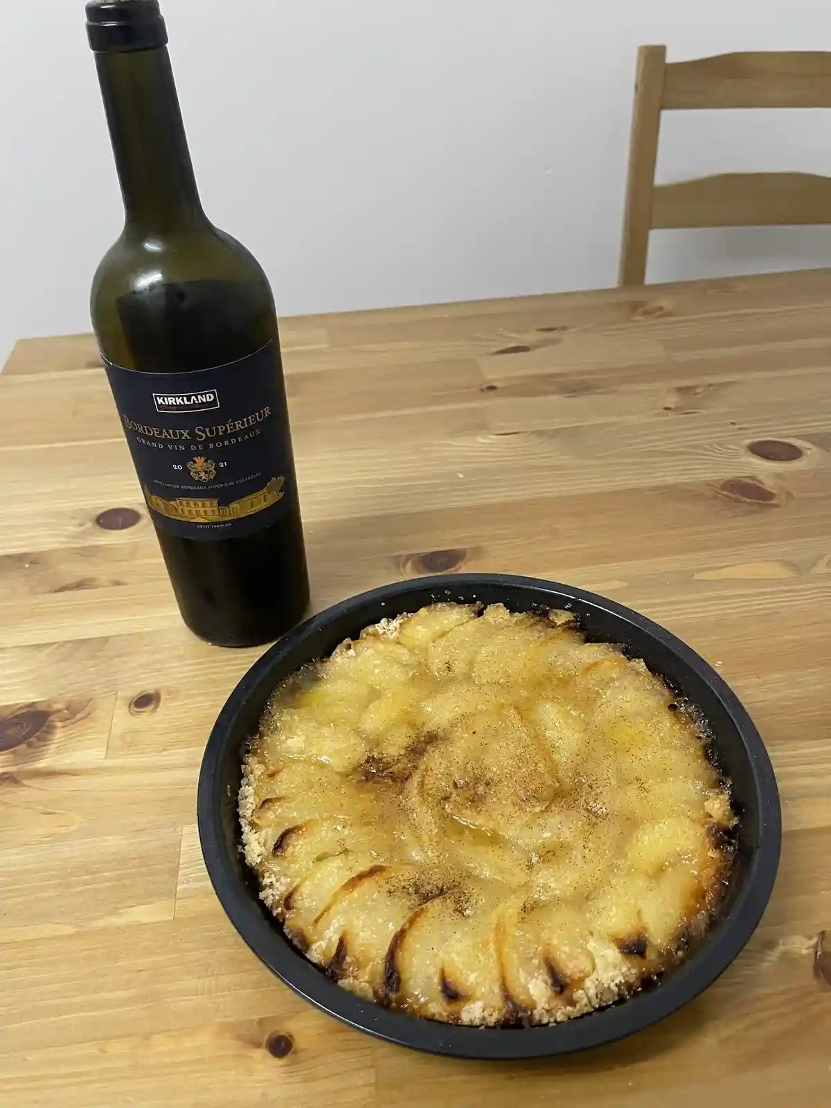 A freshly baked apple tart in a black baking dish sits on a wooden table next to a bottle of Kirkland Bordeaux Superior…