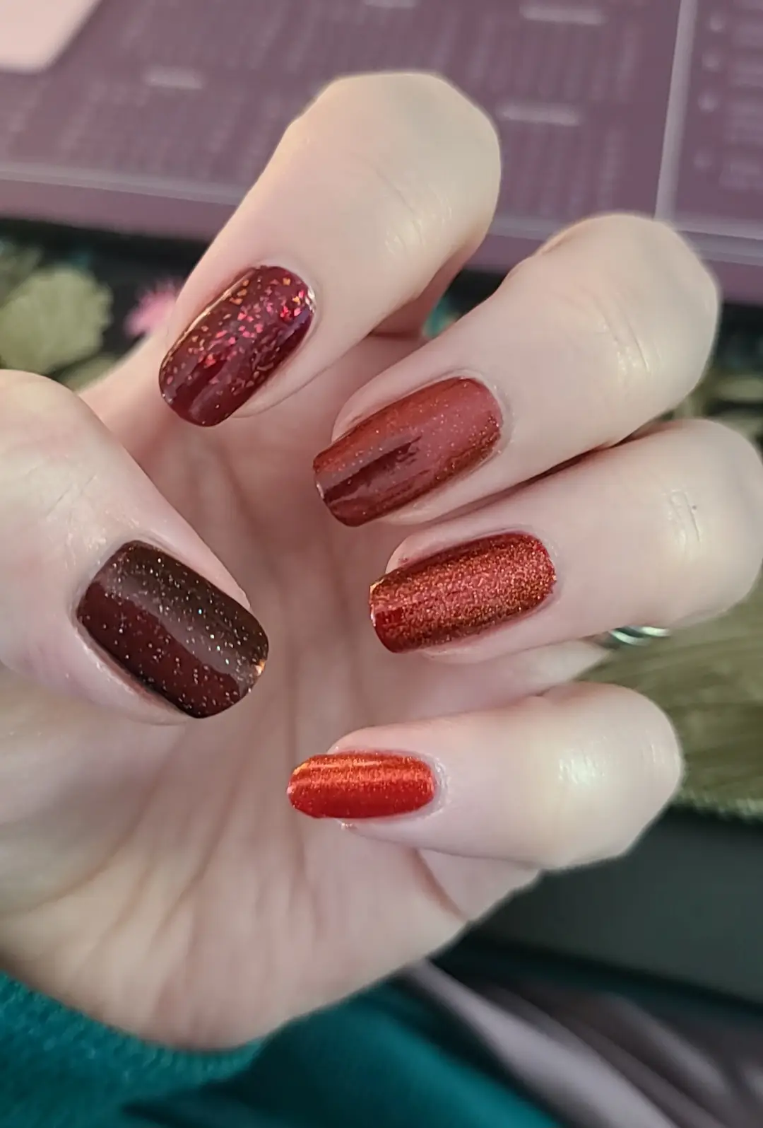 A close-up photo of a hand displaying five nails with different shades of glittery red and burgundy polish, each nail a…