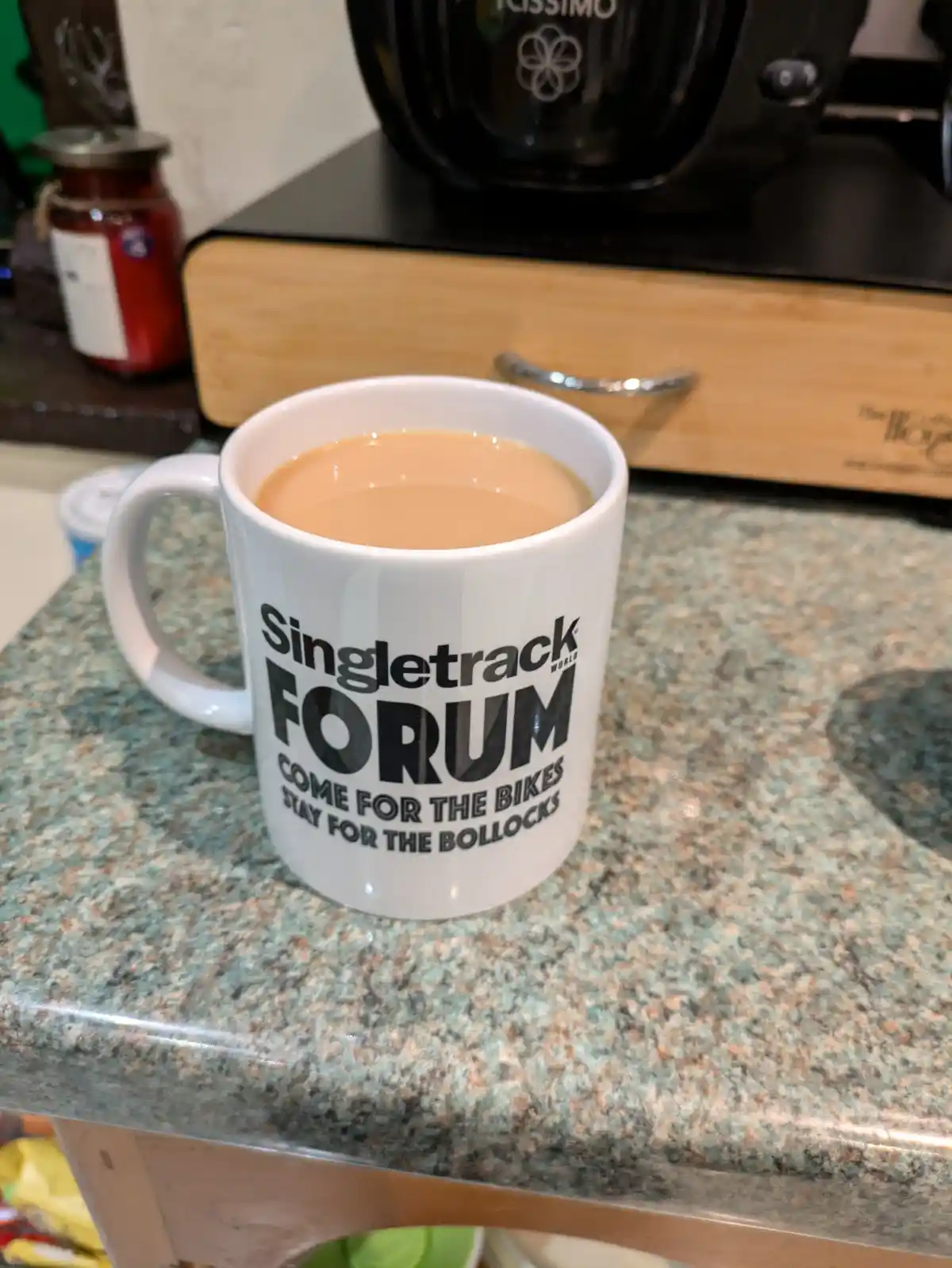 A white ceramic mug filled with light brown liquid sits on a speckled green and brown countertop. The mug has black text…