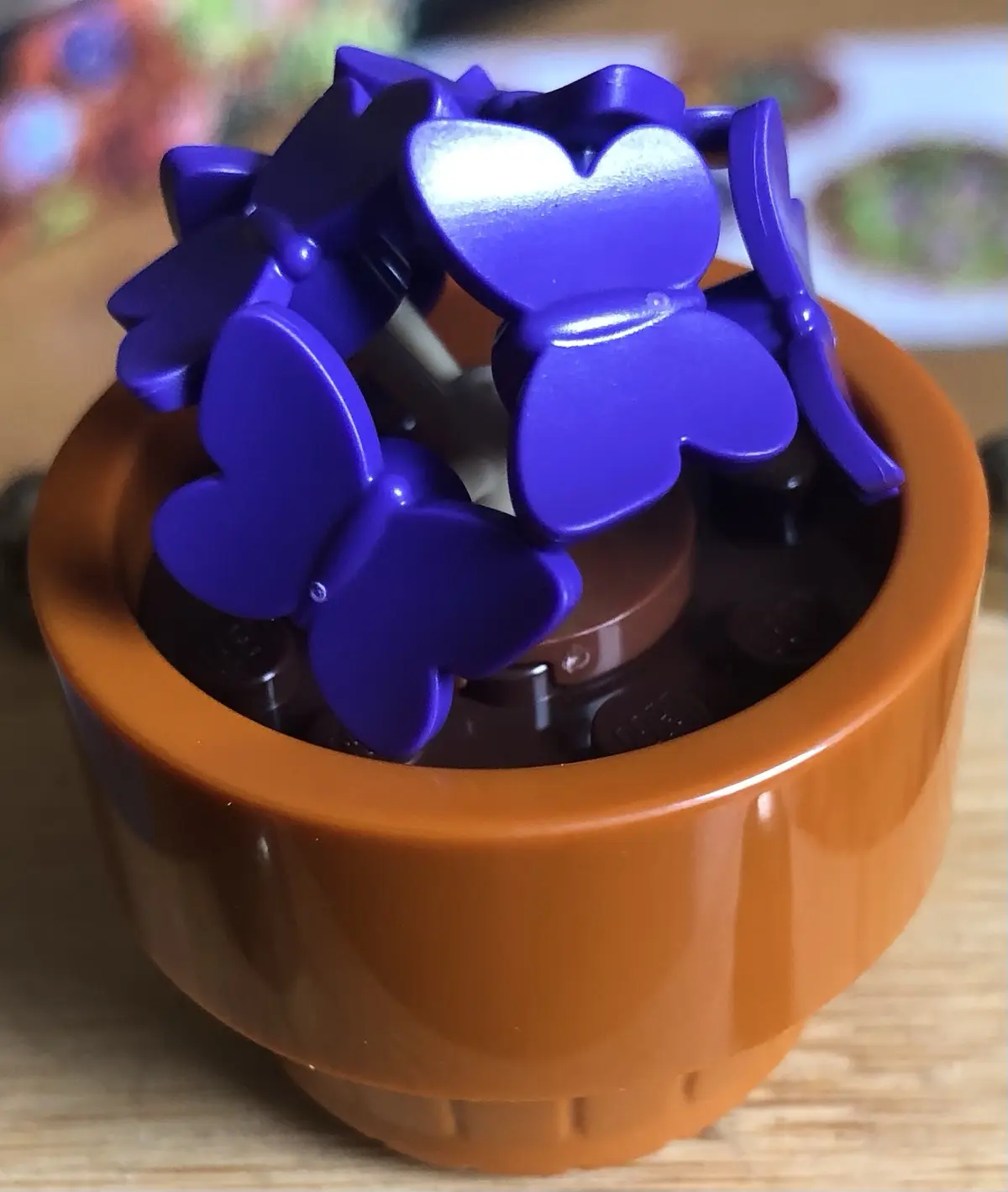 A close-up photo shows a brown plastic pot filled with dark brown LEGO bricks, topped with several shiny purple LEGO…