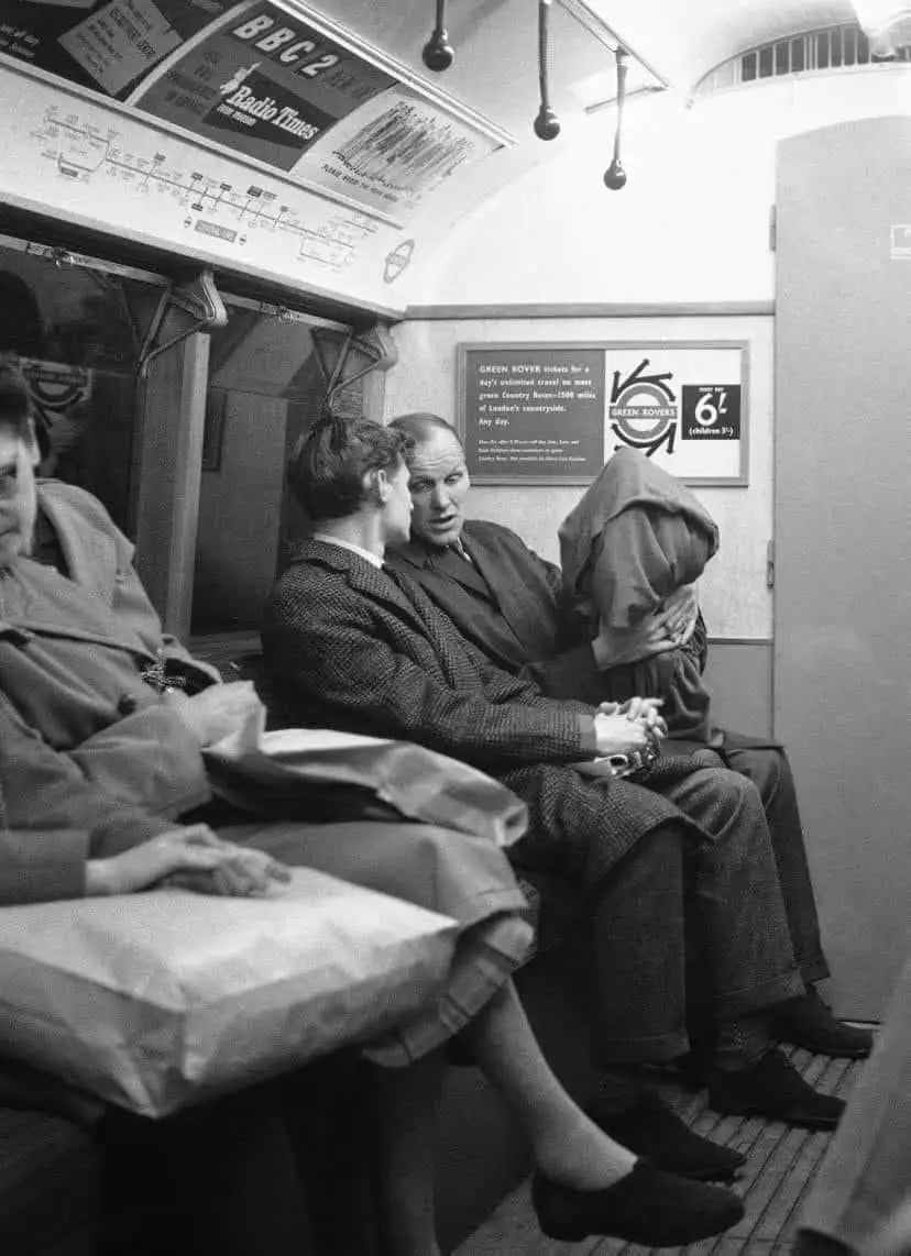 A black-and-white photo shows three people seated inside a vintage train carriage. A man in a suit is holding a bundled-up…