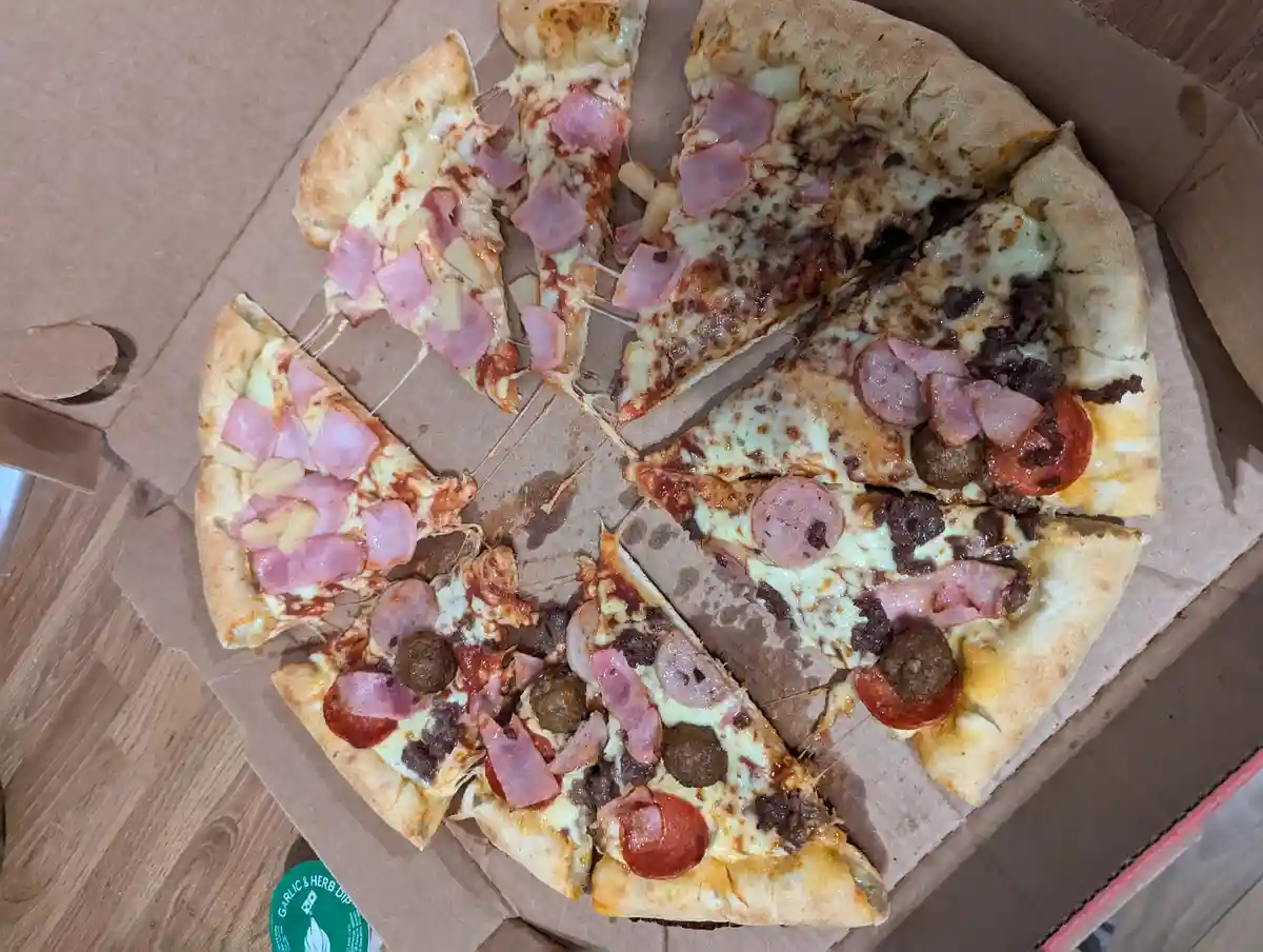An overhead photo of a pizza in a cardboard box, sliced into eight pieces, with half the slices topped with ham and…