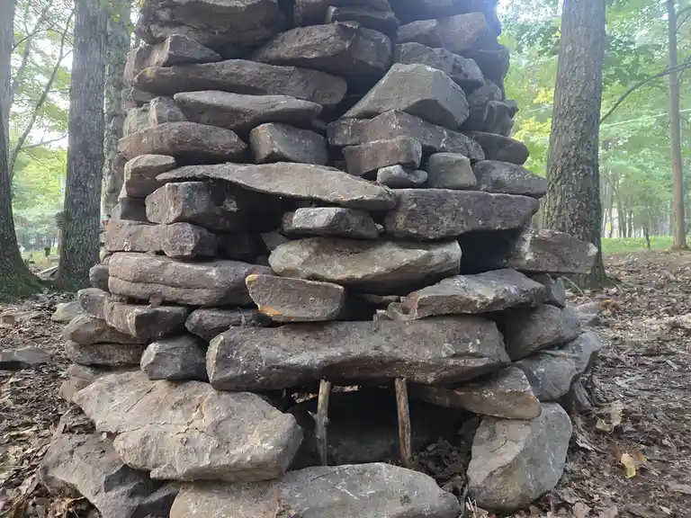 A tall, precarious stack of irregular gray stones forms a cairn in a wooded area with trees and fallen leaves on the ground…