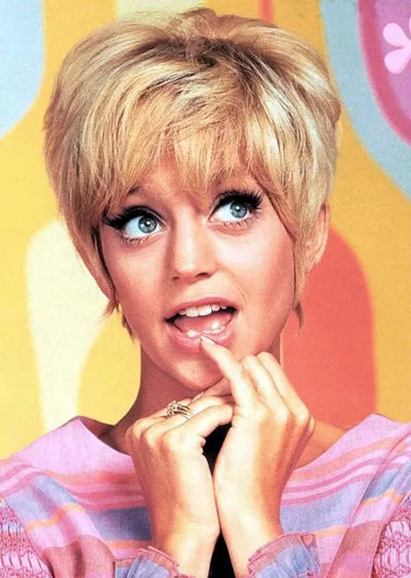 A vintage photo of a blonde woman with wide eyes and a surprised expression, her hands clasped near her mouth. She wears a…