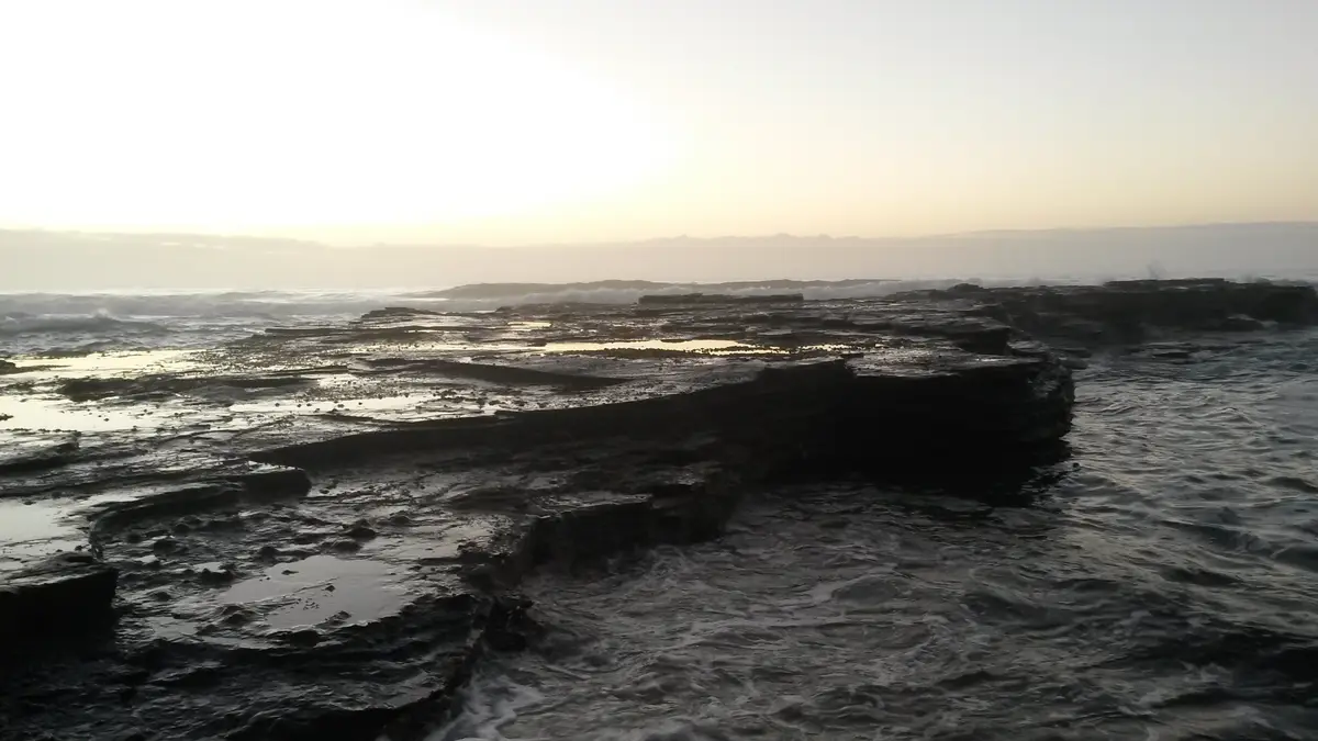 A serene, low-angle photograph of a rocky shoreline at dawn or dusk, with waves crashing against dark, wet rocks and a hazy…