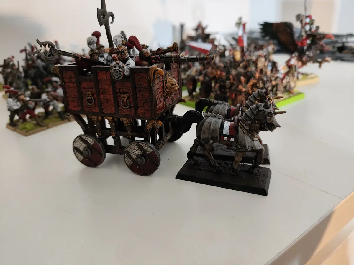 A close-up photo of a detailed miniature war wagon pulled by two armored horses, filled with armed figures, set on a white…