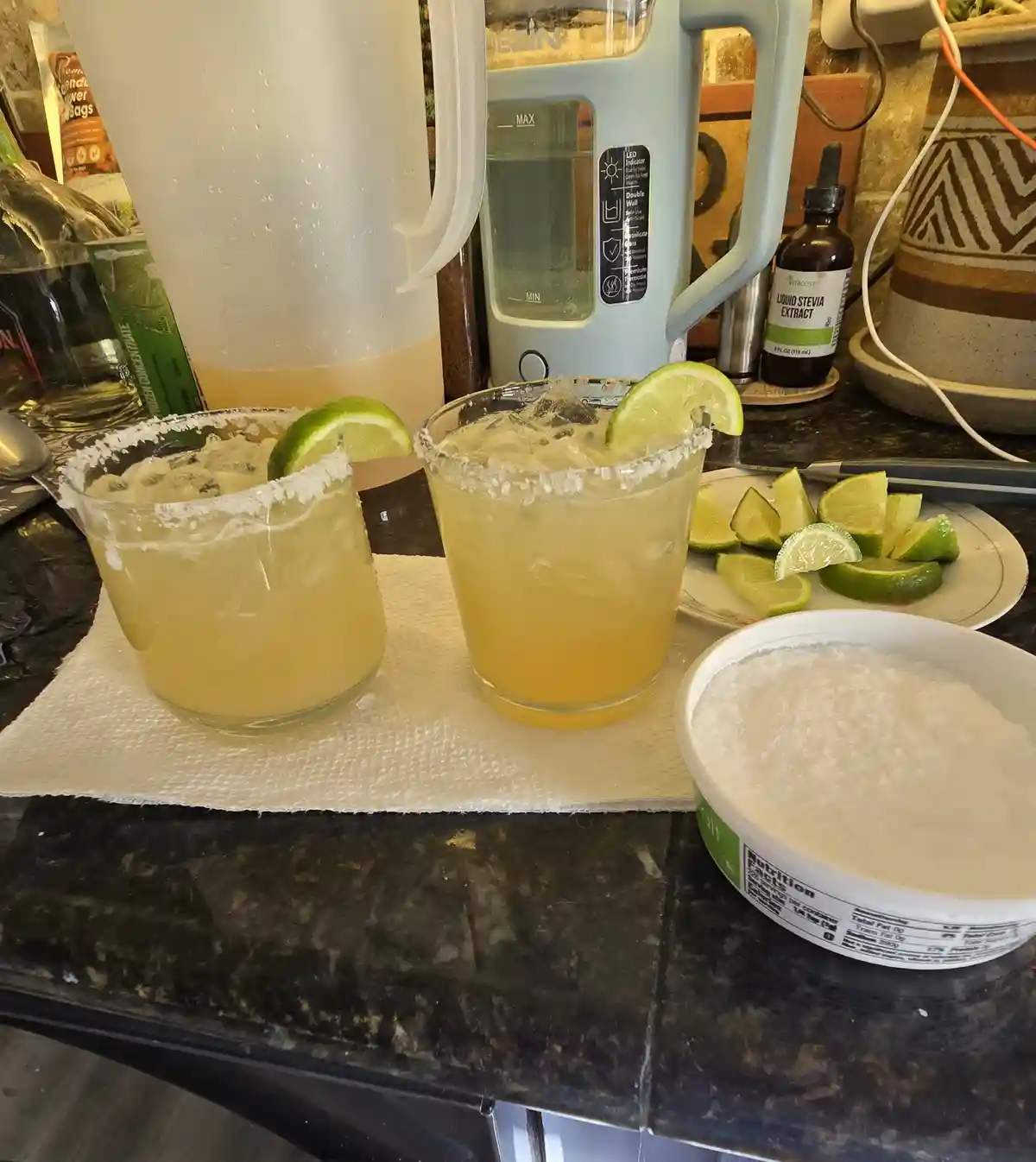 Two glasses of yellow margaritas with salt rims and lime wedges sit on a paper towel on a dark countertop, next to a bowl of…