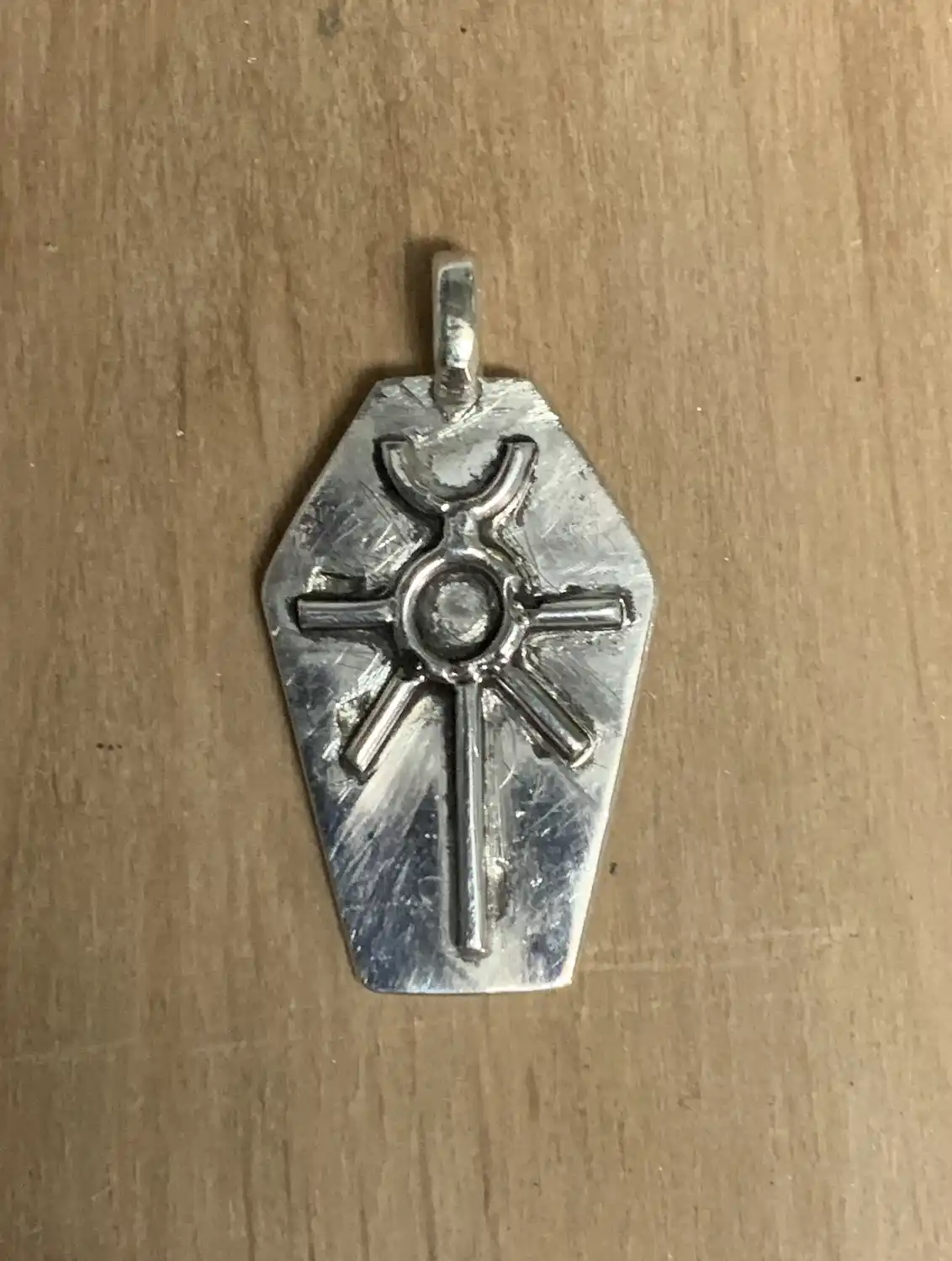 A silver, coffin-shaped pendant with a stylized sun or starburst design rests on a wooden surface. The pendant has a loop at…
