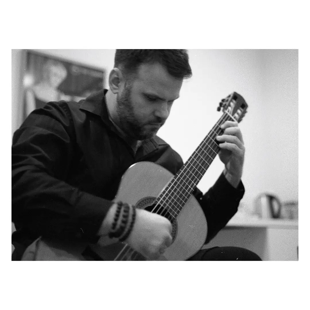 A black-and-white photo shows a man with a beard, wearing a dark shirt and beaded bracelets, intensely focused on playing a…