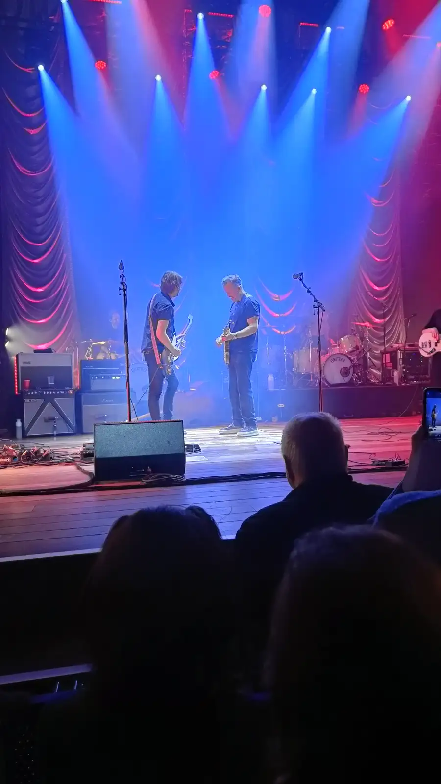 A live concert photo shows two guitarists on stage under blue and red spotlights, with a drummer and amps in the background…