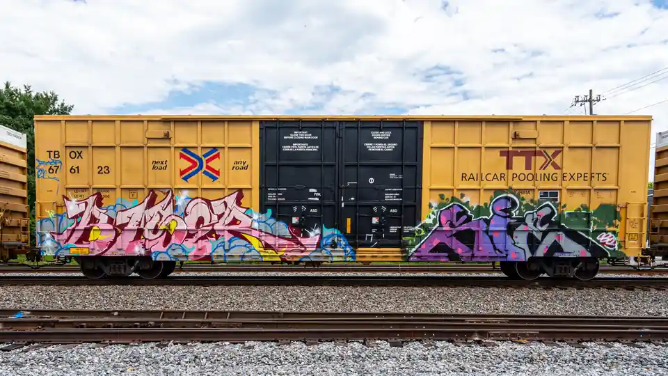 A yellow freight railcar with graffiti on its sides sits on tracks under a cloudy sky. The car has 'TTX RAILCAR POOLING…