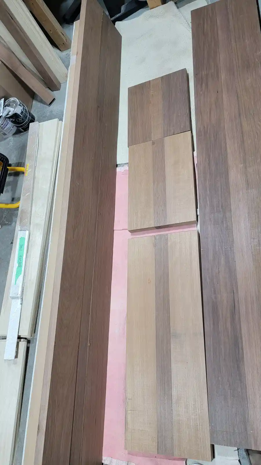 An overhead shot of a woodworking workshop showing long planks of dark and light wood arranged on a pink protective mat…