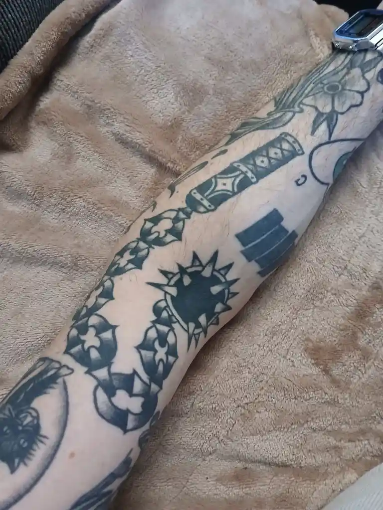 A person's forearm is covered in black-and-gray tattoos depicting various weapons and symbols from the video game 'Hollow…