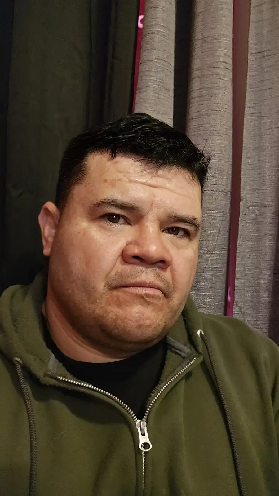A man with short dark hair and a green zip-up hoodie stares directly at the camera with a stern, unamused expression. Behind…