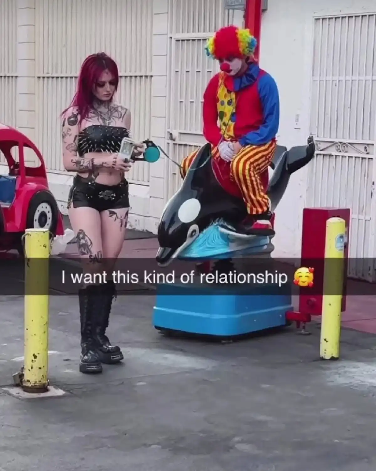A heavily tattooed woman with red hair stands beside a clown riding a mechanical orca ride, holding money and a phone. The…