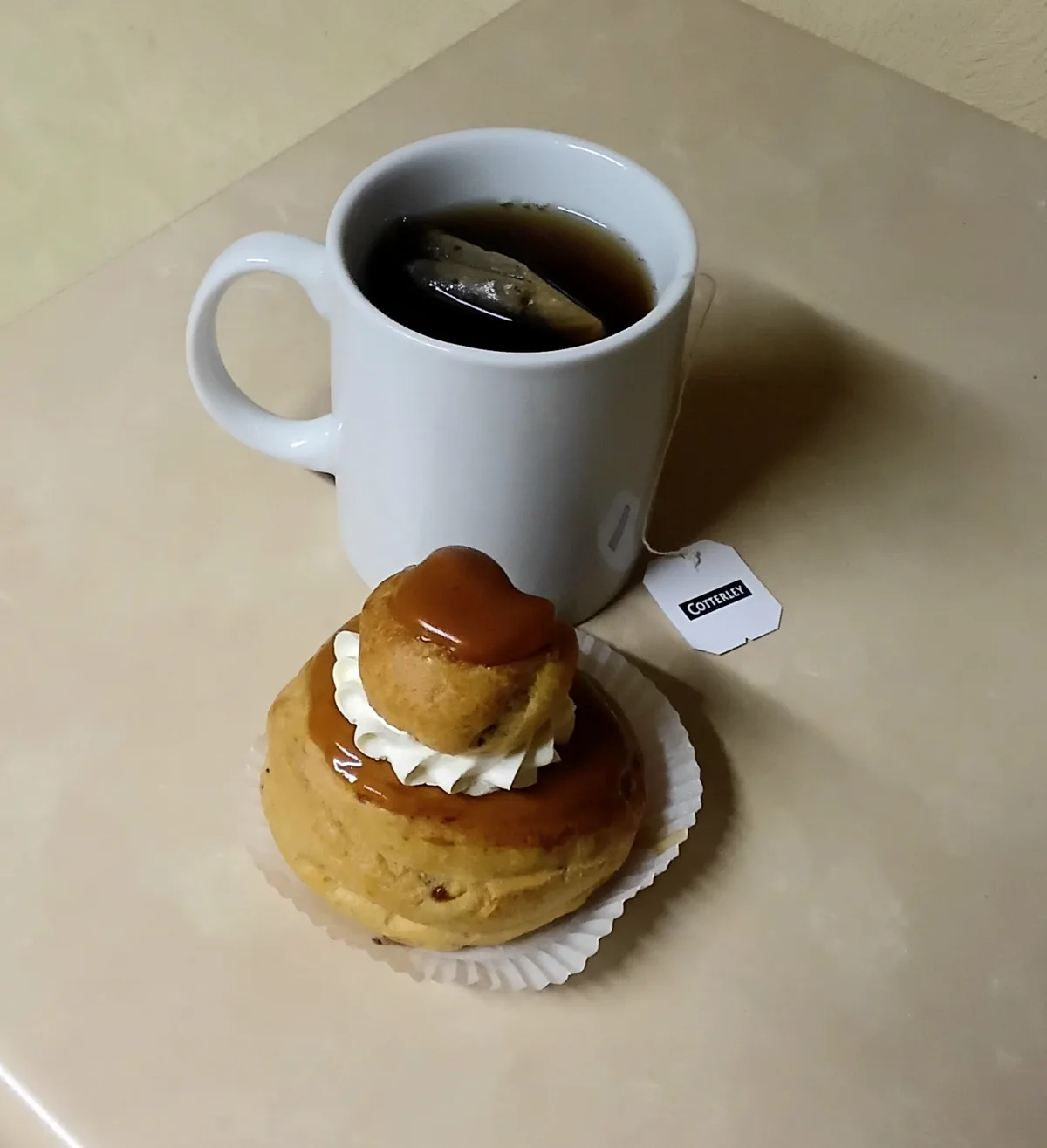 A white mug of tea with a tea bag and a 'Cotterley' tag sits beside a caramel-topped pastry with whipped cream on a paper…