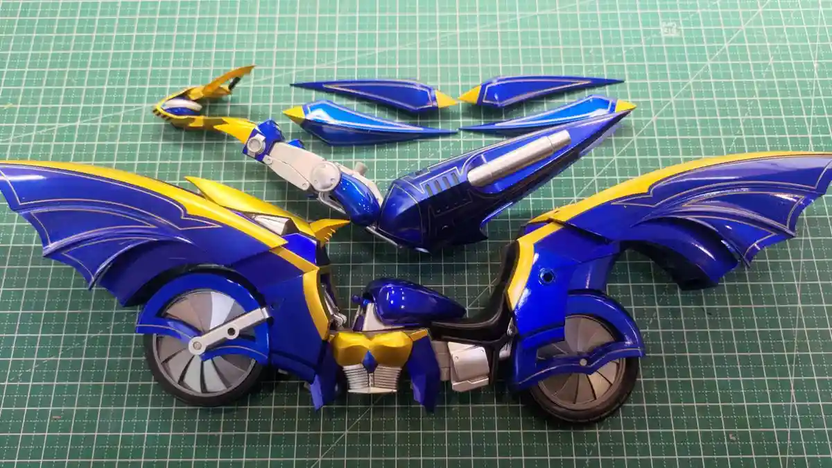 A blue and yellow transforming toy motorcycle is partially assembled on a green cutting mat, with its wings and other parts…