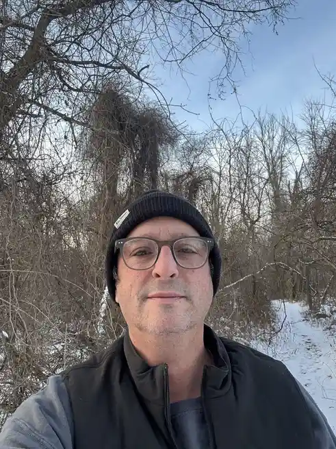 A man wearing glasses and a black beanie takes a selfie outdoors in a snowy, wooded area with bare trees. Behind him, a…