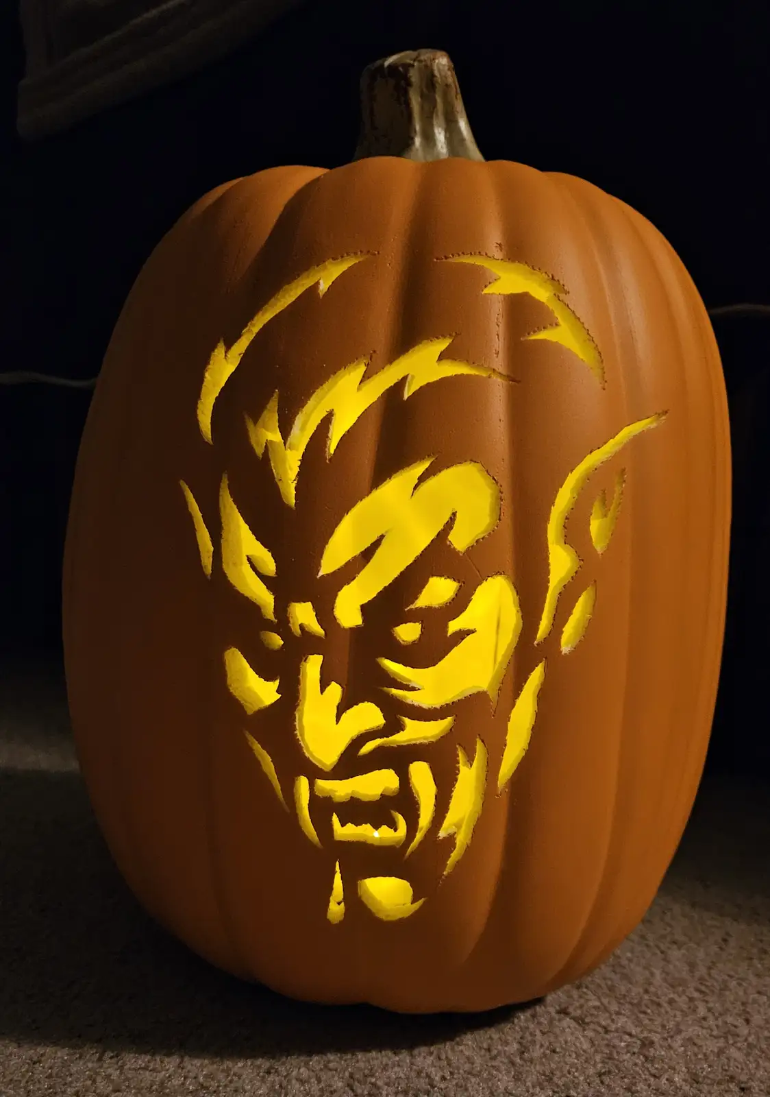 A close-up of an orange pumpkin carved with a detailed, menacing demonic face, illuminated from within by a warm yellow…