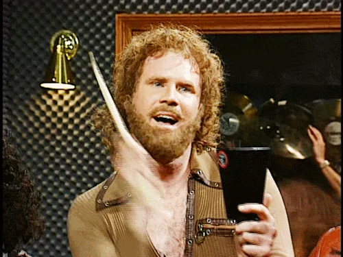 A man with curly hair and a beard, wearing a brown shirt, holds a black cup and raises a drumstick in a celebratory or…