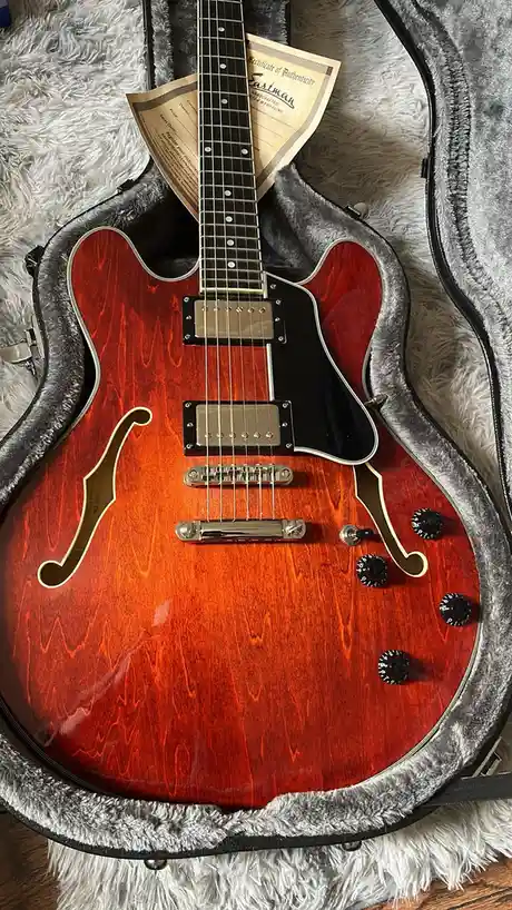 A close-up photo shows a glossy, reddish-orange Eastman semi-hollow body electric guitar resting inside its grey plush-lined…