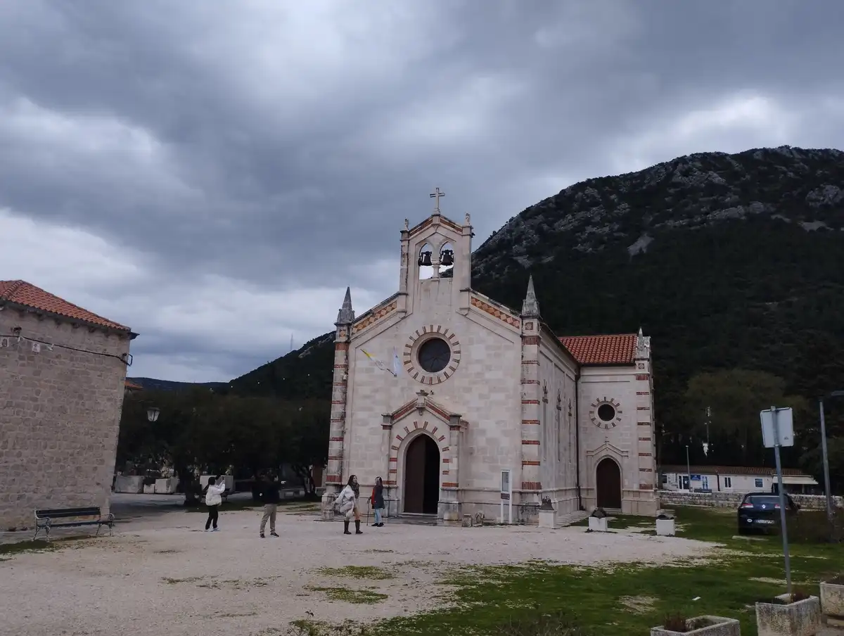 A wide shot of a stone church with a bell tower and cross under a cloudy sky, with several people in the foreground. The…