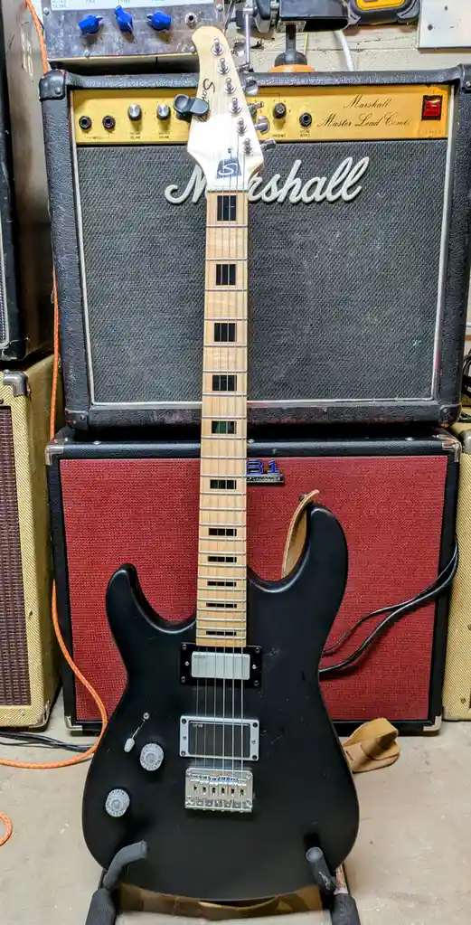A black electric guitar with a light wood neck and EMG pickups is propped up in front of two stacked Marshall amplifiers…