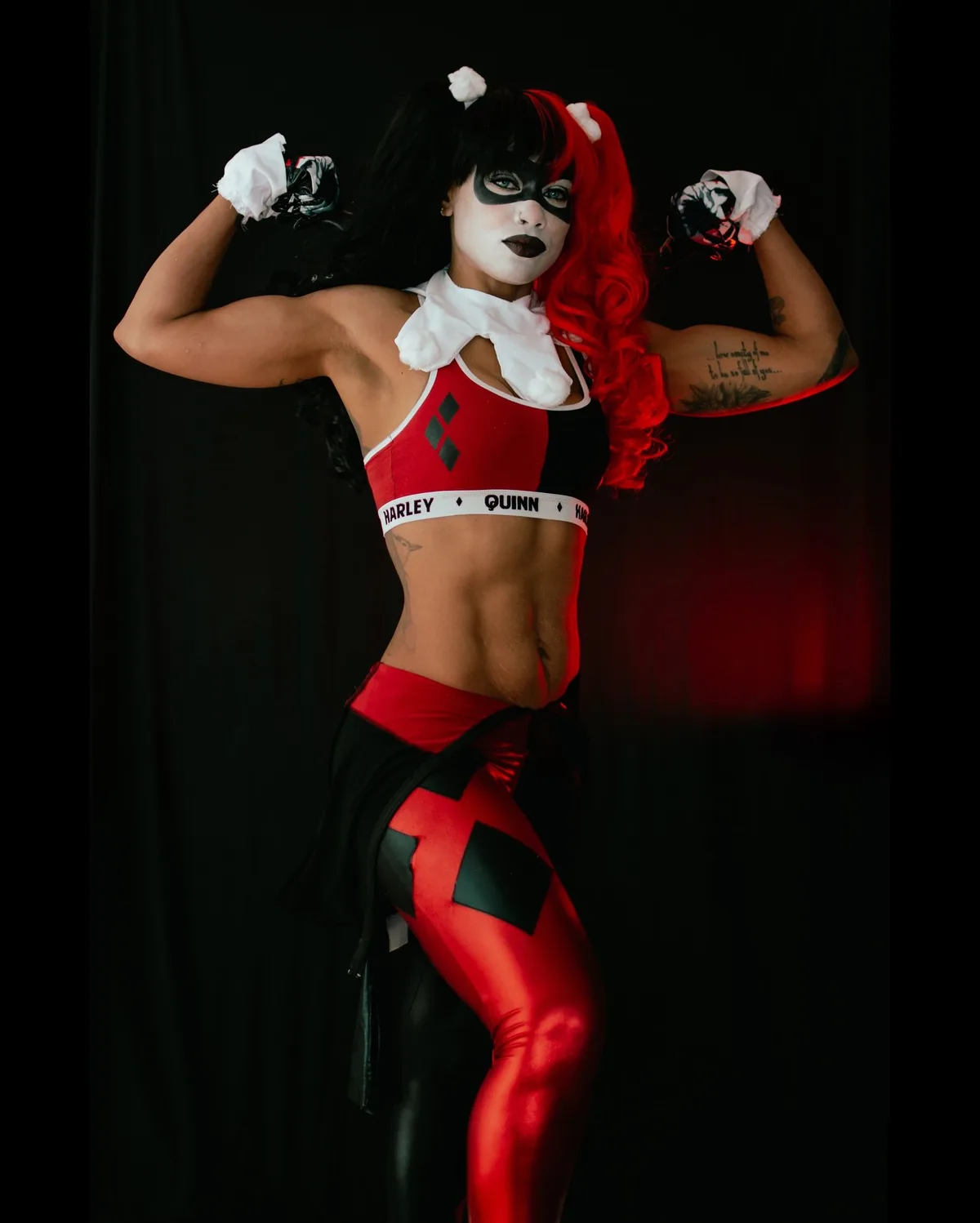 A muscular woman in a Harley Quinn cosplay, flexing her biceps against a black background. She wears a red and black outfit…