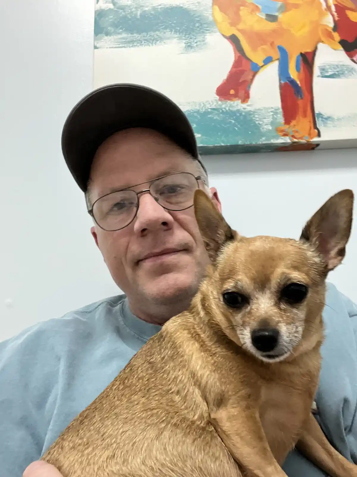 A man wearing glasses and a black cap holds a small tan dog in front of him; behind them is a colorful abstract painting of…