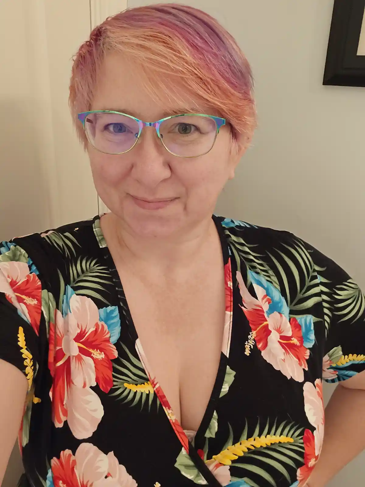A person with short, multi-toned pink and peach hair and iridescent glasses wears a black wrap dress with a bold tropical…