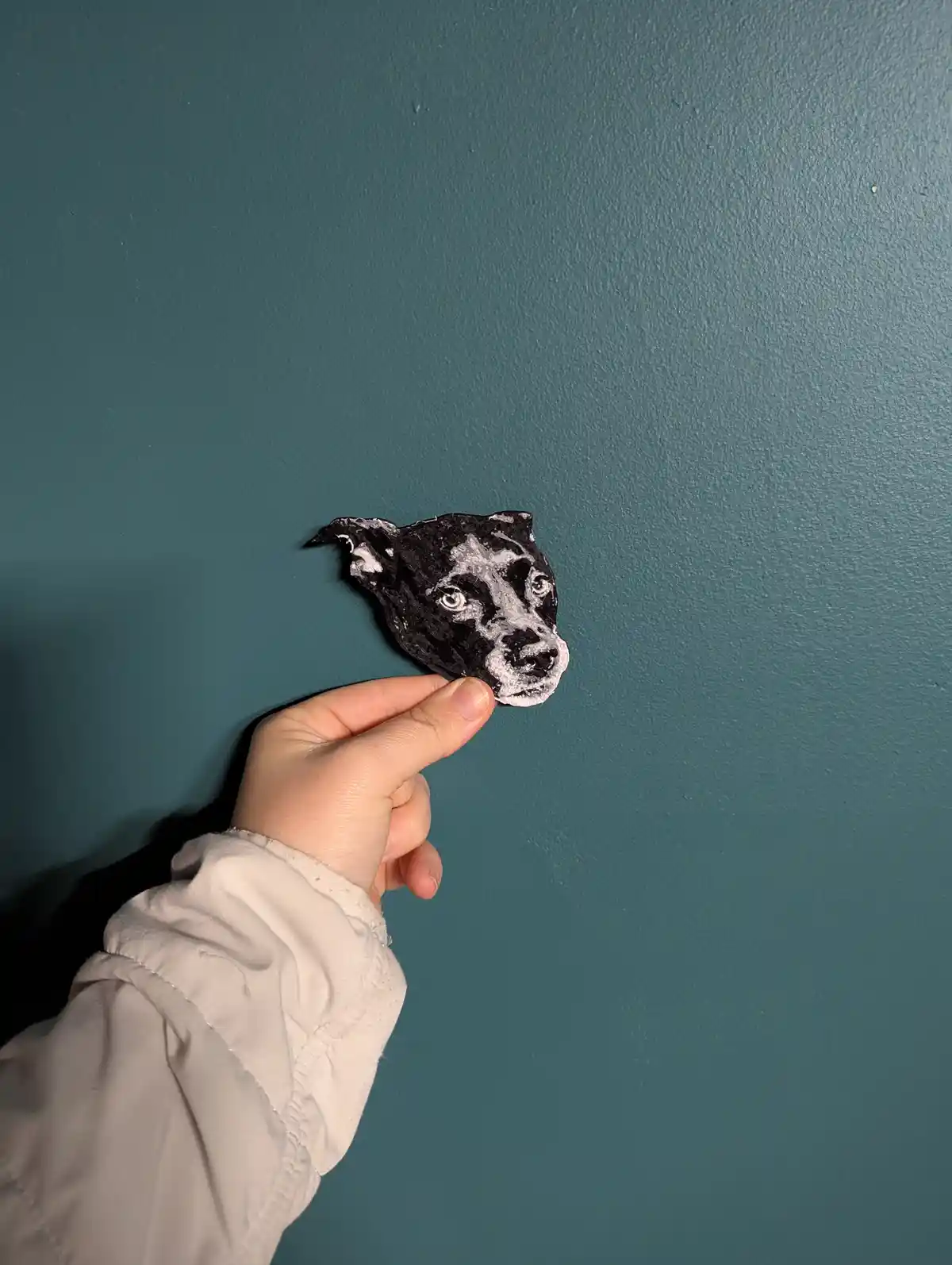 A hand holds up a small, black-and-white painted cutout of a dog's face against a teal wall. The text overlaid on the image…