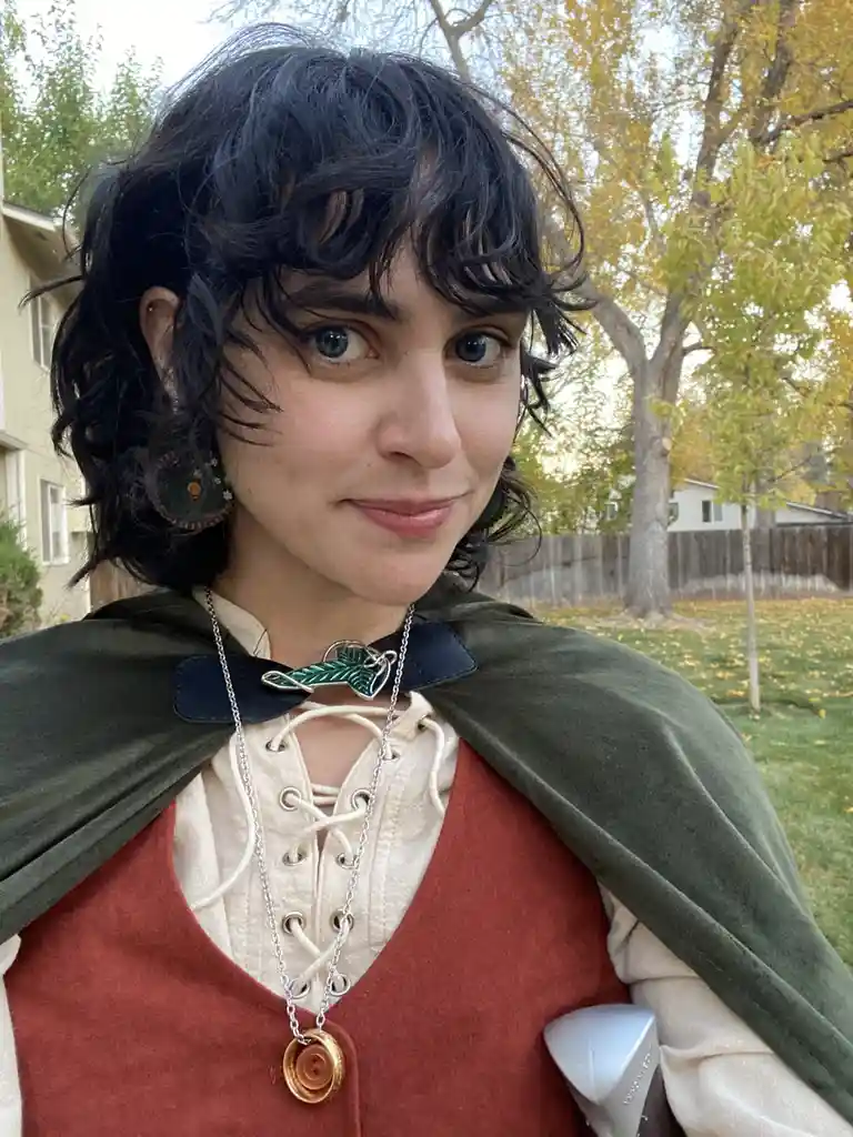 A person with dark curly hair and blue eyes wears a fantasy-style outfit including a green cloak, a red vest over a laced…