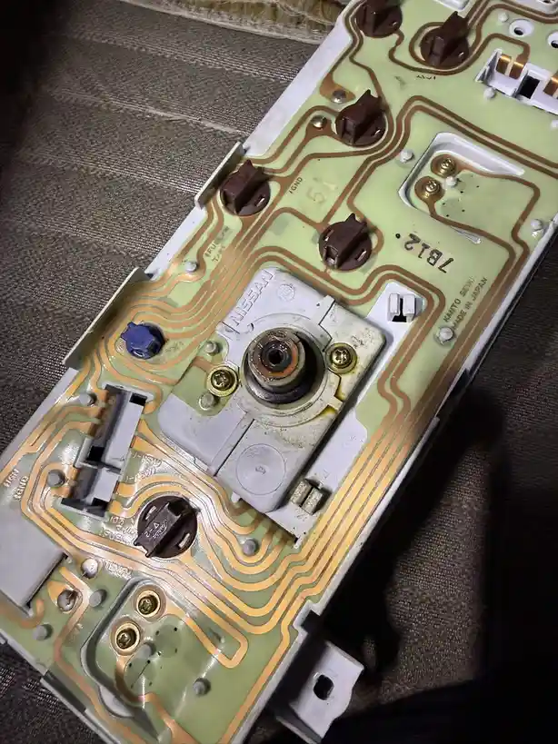 A close-up photo of a green circuit board from a Nissan 7B12 engine control unit, showing copper traces, connectors, and a…