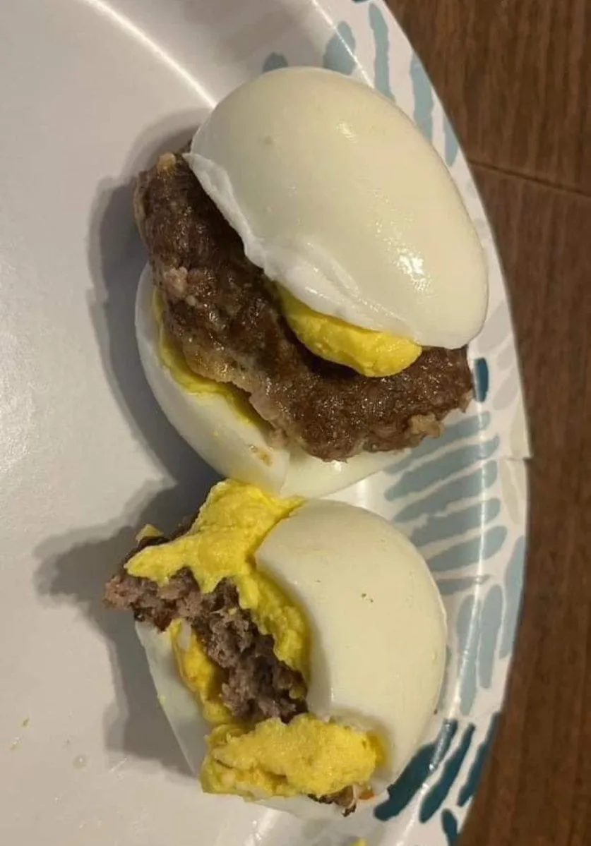 two halves of a hard-boiled egg are used as buns for a meat patty, with one half intact and the other half broken open to…