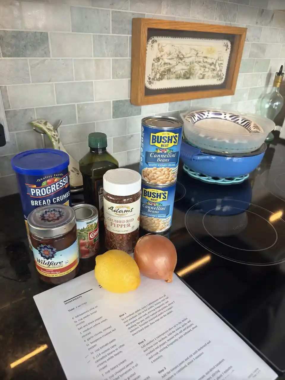 A kitchen counter displays ingredients for a recipe, including two cans of Bush's Best Cannellini Beans, a lemon, an onion…