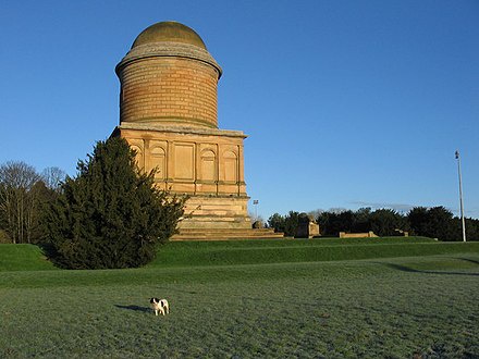 A small dog stands in a grassy field in front of a large, domed stone mausoleum under a clear blue sky. The image has text…