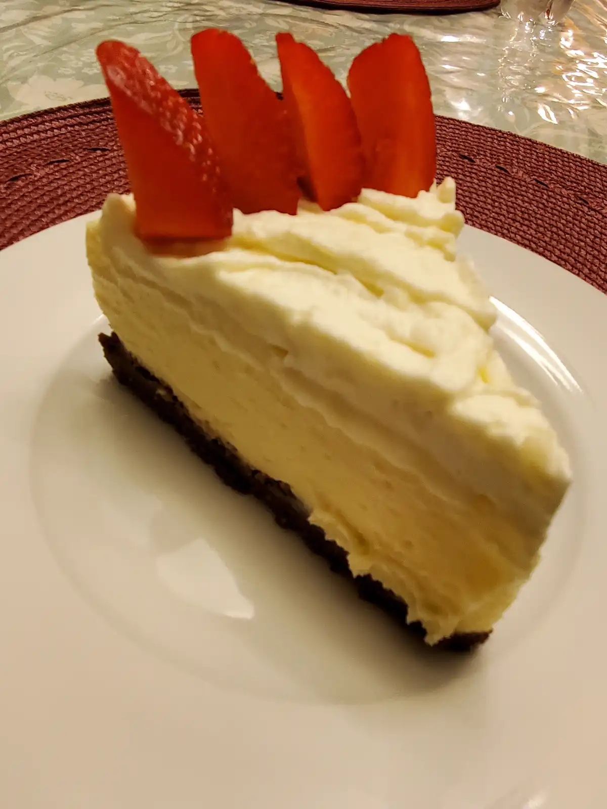 A slice of creamy cheesecake with a dark crust sits on a white plate, topped with four red strawberry slices arranged like a…