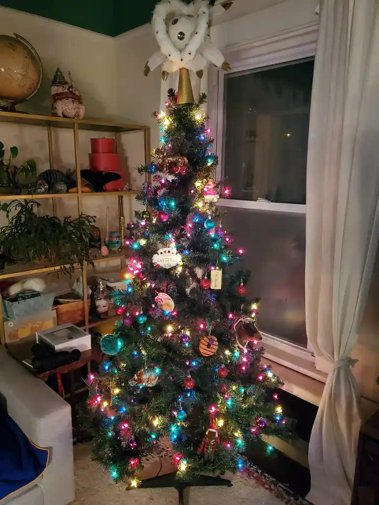 A brightly lit, decorated christmas tree with colorful lights and various ornaments stands in a living room, featuring a…