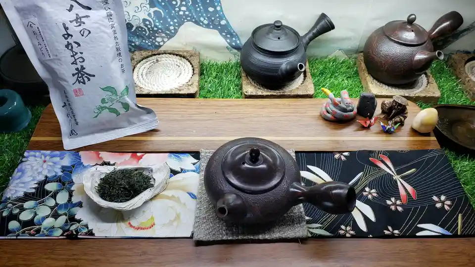 A serene Japanese tea setup features a dark ceramic teapot on a woven mat, beside a shell holding green tea leaves, with a…
