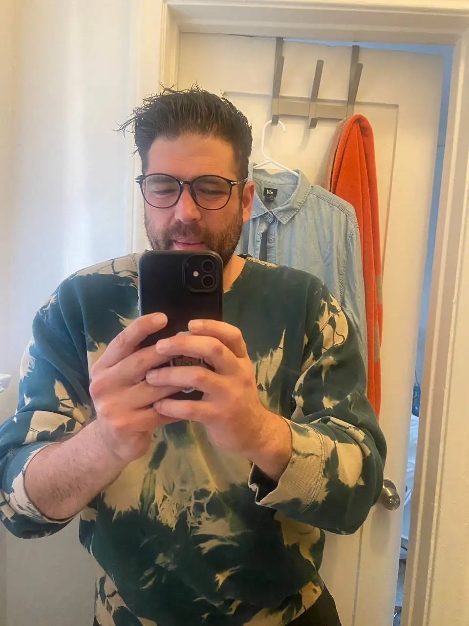 A man with glasses and a beard takes a mirror selfie holding a black smartphone, wearing a green and beige tie-dye…