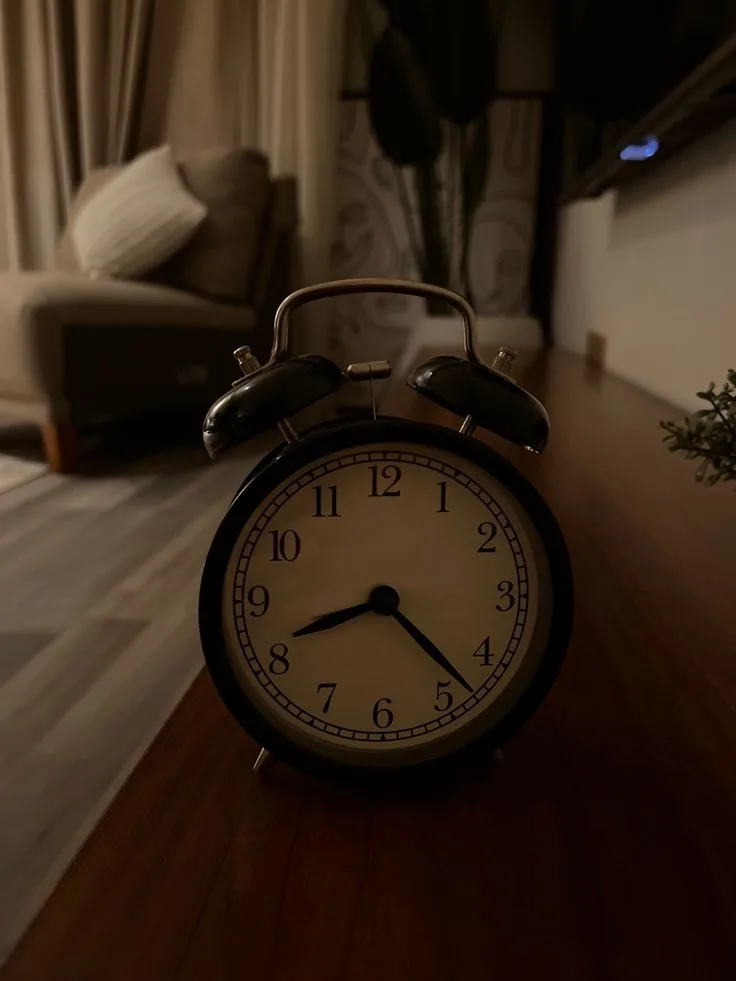 A black, classic twin-bell alarm clock sits on a wooden surface in a dimly lit room, its hands pointing to 4:20. The…