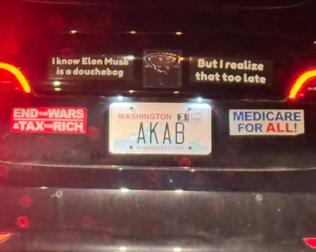 The rear of a dark car at night shows a Washington license plate with the letters AKAB. Above the plate are two stickers…