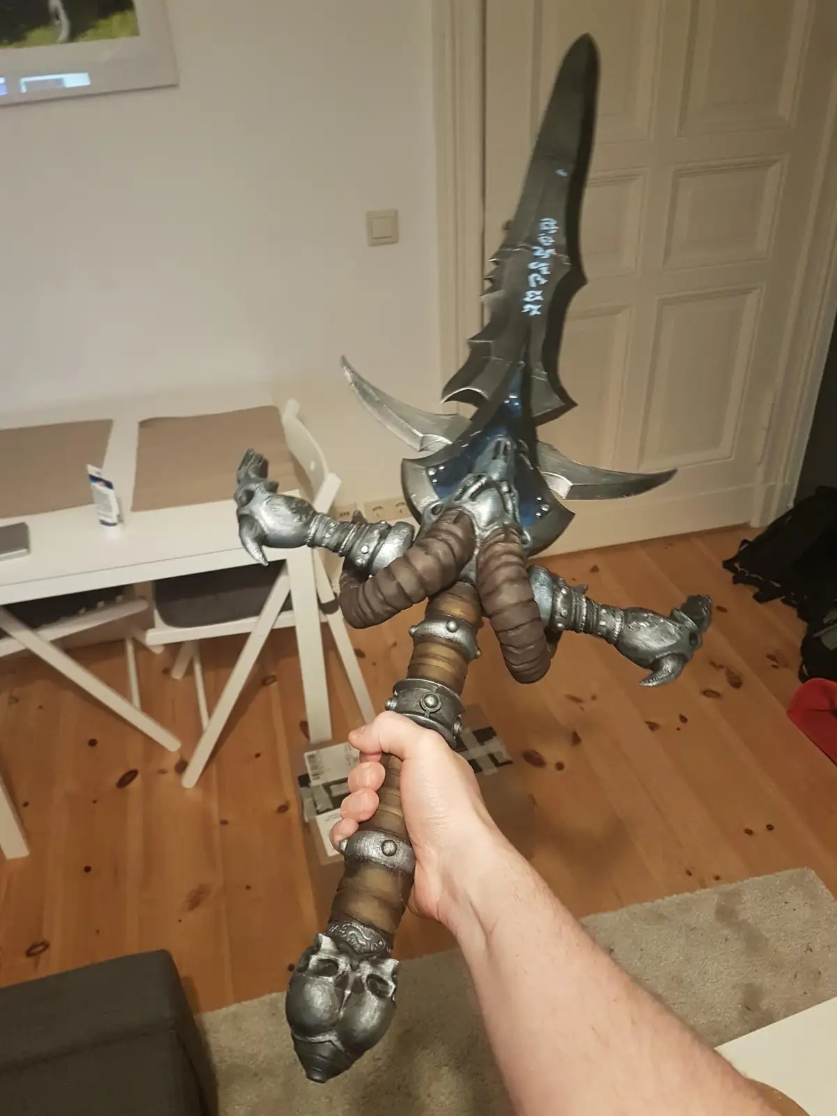 A person holds a large, ornate fantasy sword with multiple blades and claw-like grips in a domestic room with wooden floors…