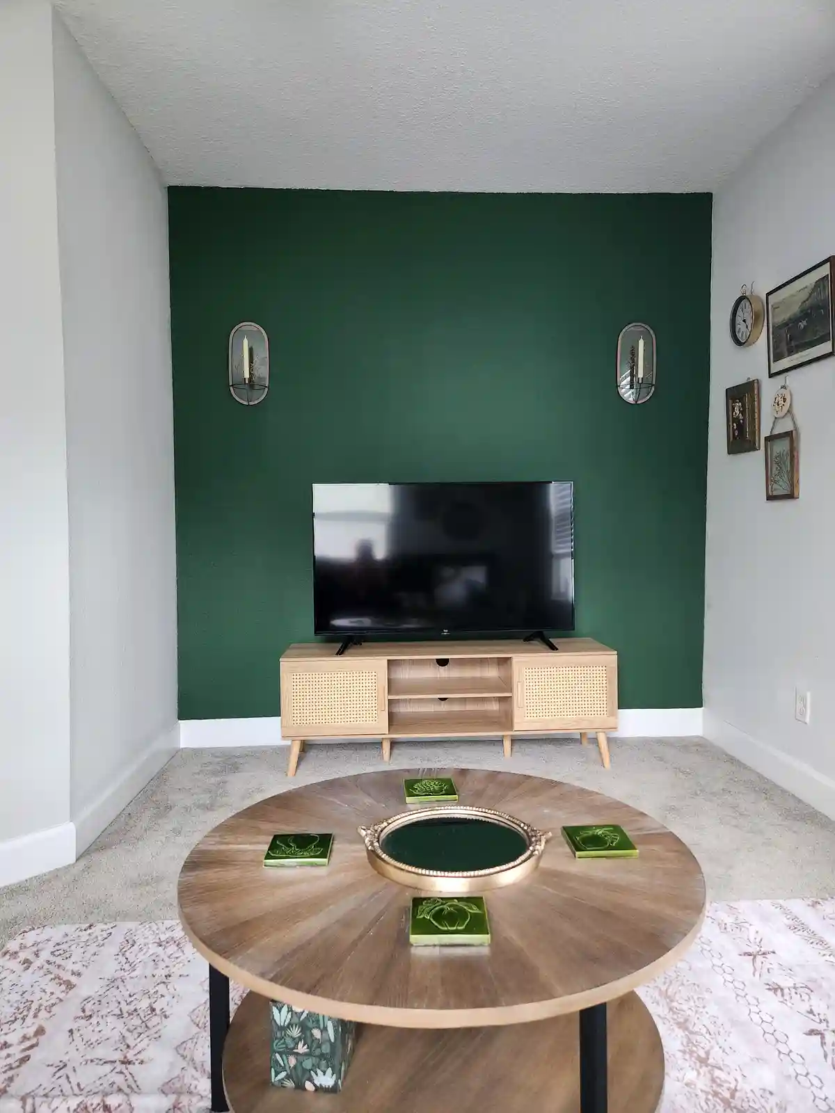 A living room features a dark green accent wall behind a TV on a light wood media console. A round wooden coffee table in…