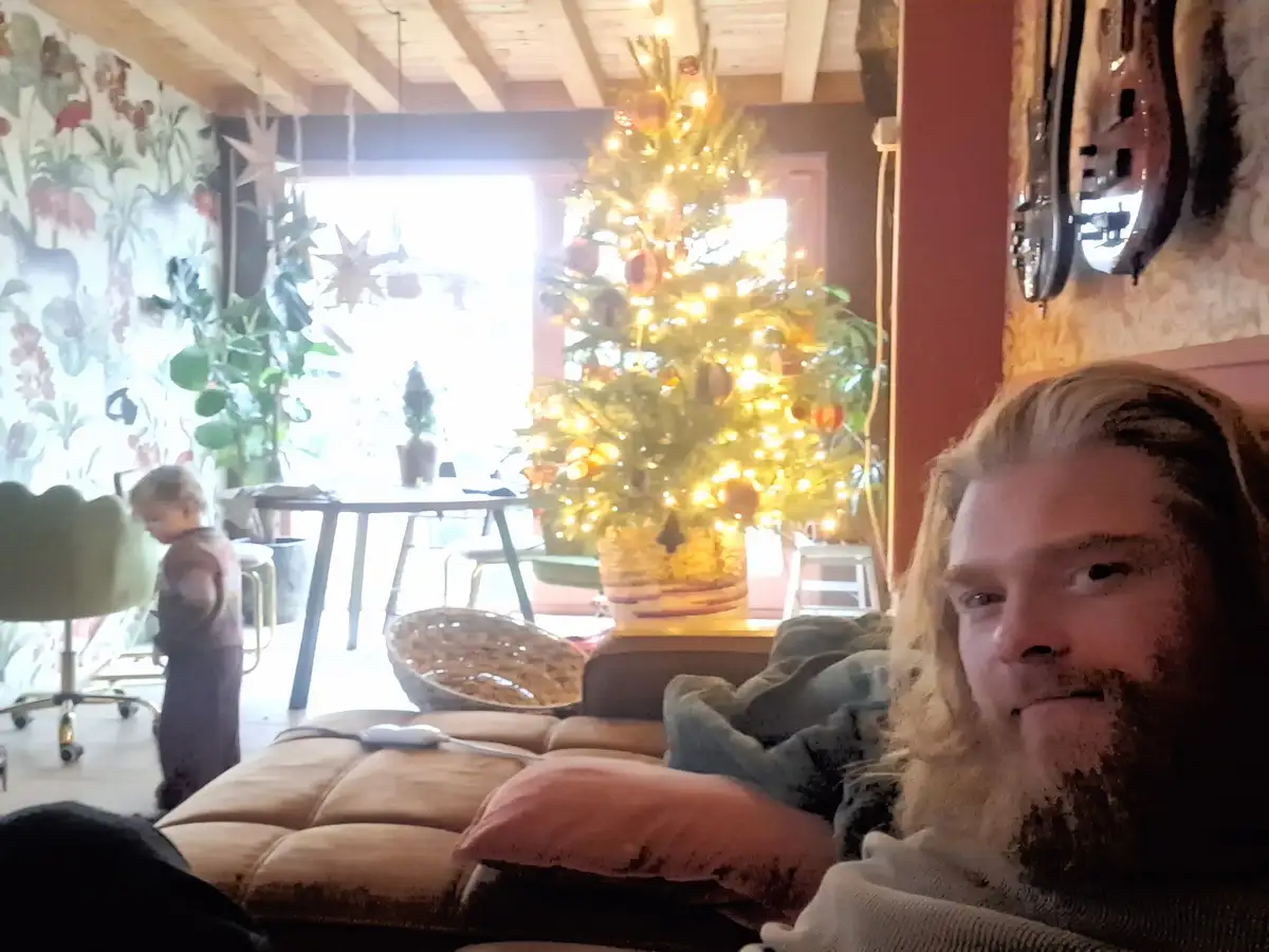 A man with long blonde hair and a beard lies on a couch, looking directly at the camera with a relaxed expression. Behind…